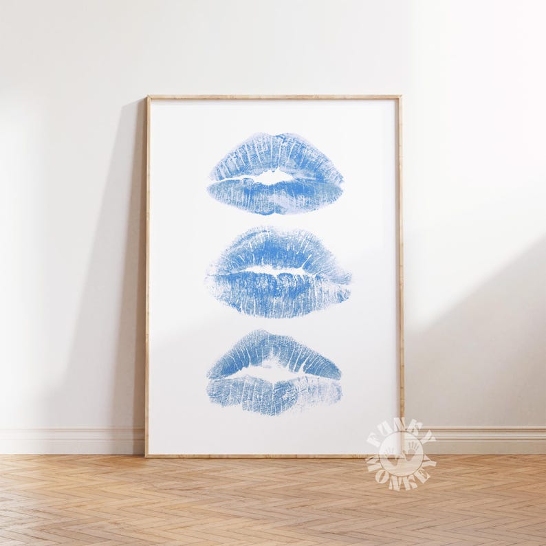 May include: A framed print featuring three blue lipstick kiss marks on a white background. The artwork is displayed in a light-colored wooden frame and rests on a wooden floor.