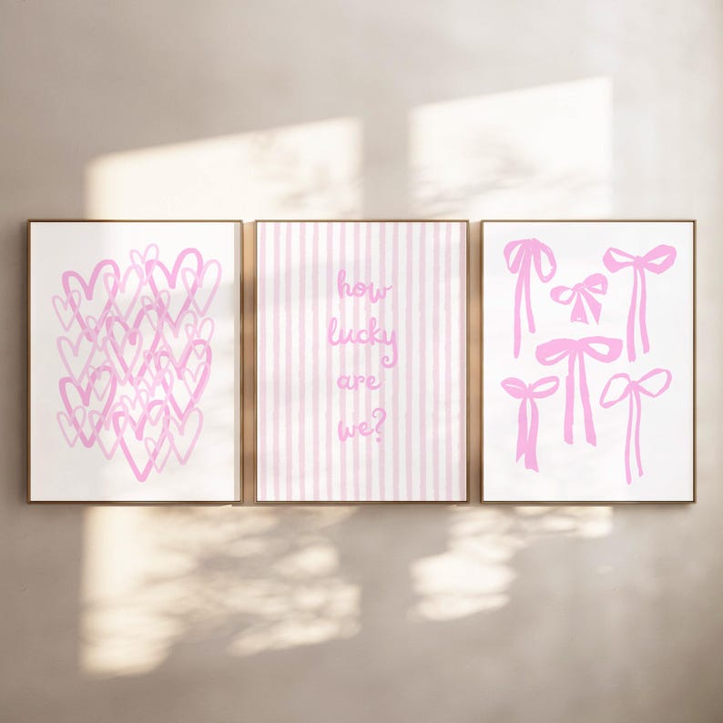 May include: Three framed art prints with a light pink and white color scheme. The left print features pink hearts, the center print has pink stripes and the text "how lucky are we?", and the right print shows pink bows.
