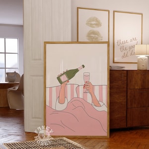 K&ouml;nnte beinhalten: Ein gerahmter Kunstdruck zeigt eine Person, die im Bett Champagner in ein Glas gie&szlig;t. Das Kunstwerk zeigt einen rosa-wei&szlig; gestreiften Hintergrund, eine rosa Decke und eine gr&uuml;ne Champagnerflasche. Zwei weitere gerahmte Drucke sind im Hintergrund zu sehen.