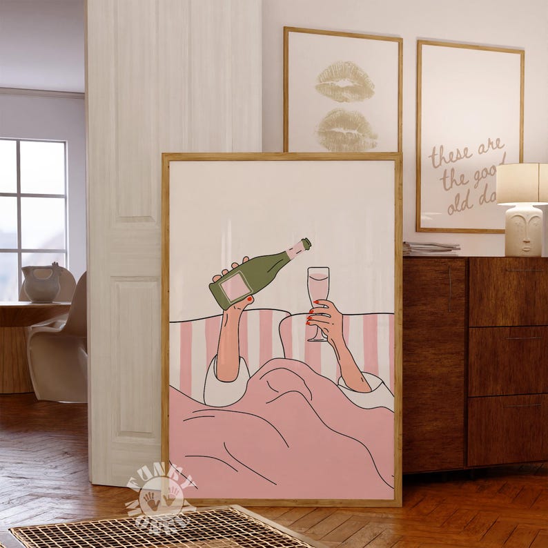 K&ouml;nnte beinhalten: Ein gerahmter Kunstdruck zeigt eine Person, die im Bett Champagner in ein Glas gie&szlig;t. Das Kunstwerk zeigt einen rosa-wei&szlig; gestreiften Hintergrund, eine rosa Decke und eine gr&uuml;ne Champagnerflasche. Zwei weitere gerahmte Drucke sind im Hintergrund zu sehen.