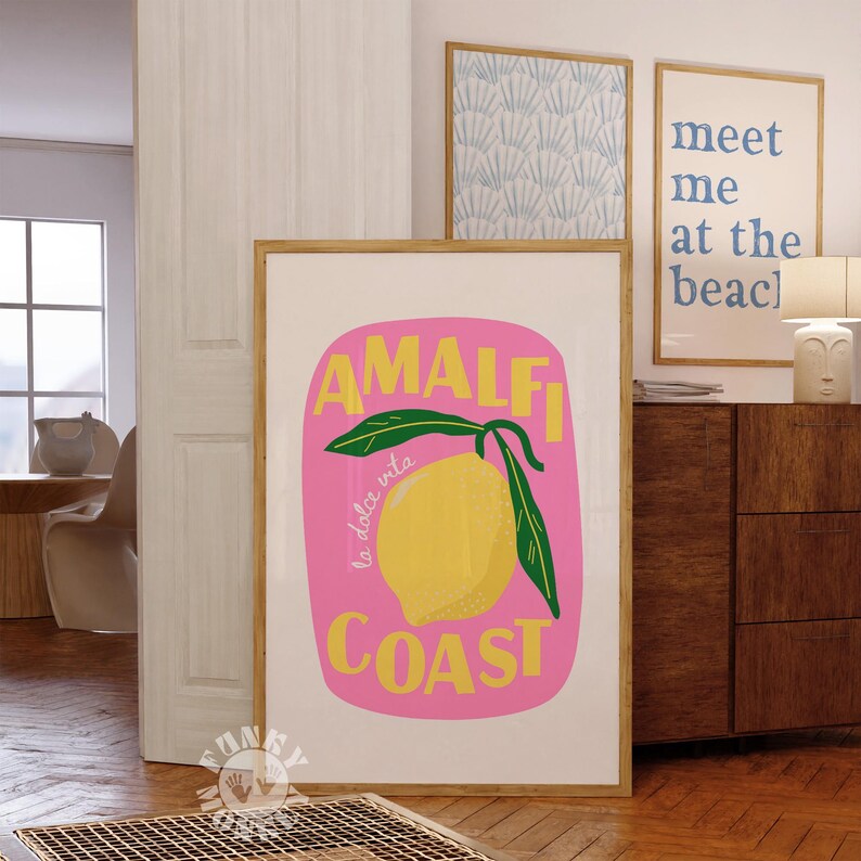 May include: A framed art print featuring a vibrant illustration of a lemon with the words "AMALFI COAST" in yellow and green on a pink background. The phrase "la dolce vita" is also included. Additional framed art pieces are visible.