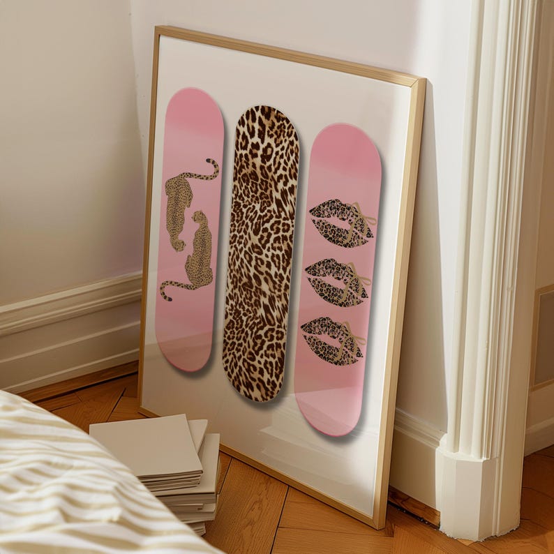Op de afbeelding: Een ingelijst kunstwerk met drie roze skateboard decks. E&eacute;n deck heeft een luipaardprint, een ander luipaardillustraties en de derde lipprintontwerpen. Het frame is goud.