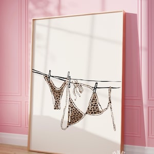 May include: A framed print of a leopard print bikini hanging on a clothesline. The print is on a white background and the frame is gold.