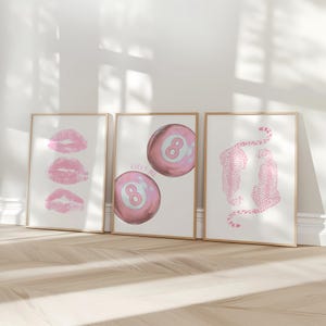 May include: Three framed art prints with a light wood finish. The prints feature pink lipstick kisses, a pink eight ball with the text "LUCKY ME", and two pink cheetahs. The prints are displayed on a white wall.