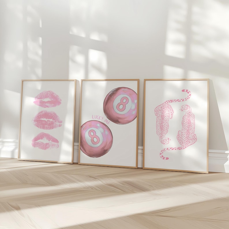 May include: Three framed art prints with a light wood finish. The prints feature pink lipstick kisses, a pink eight ball with the text "LUCKY ME", and two pink cheetahs. The prints are displayed on a white wall.