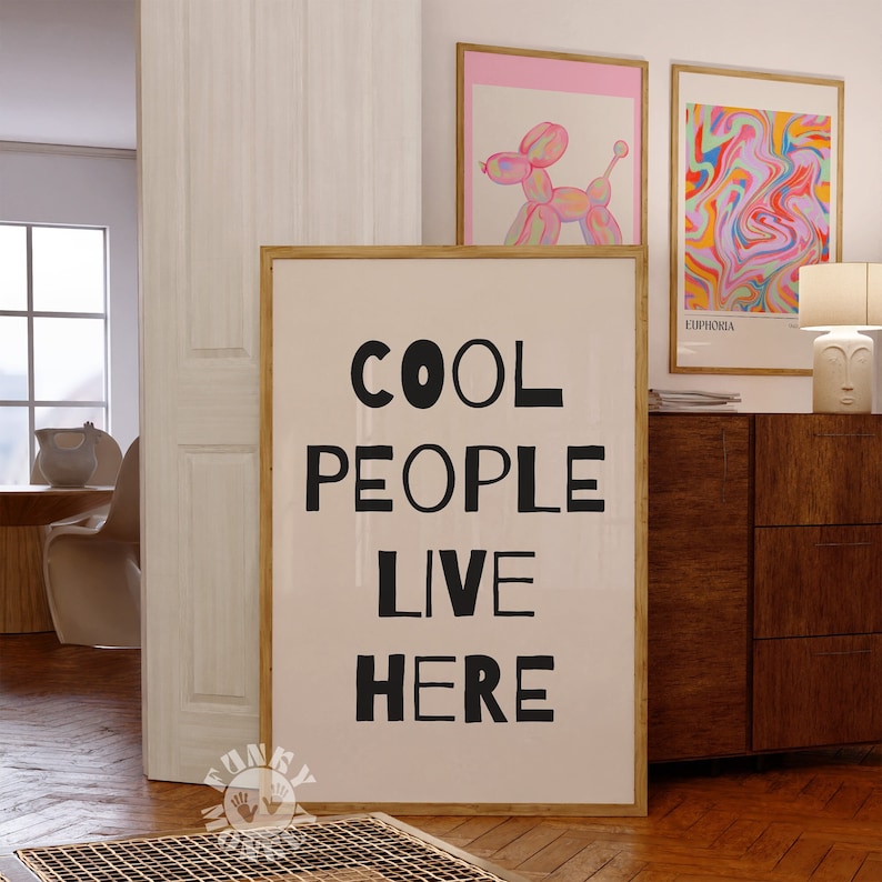 Op de afbeelding: Een ingelijst print met een witte achtergrond en zwarte tekst die "COOL PEOPLE LIVE HERE" luidt. De print hangt aan een muur in een kamer met een houten vloer en een houten kast.