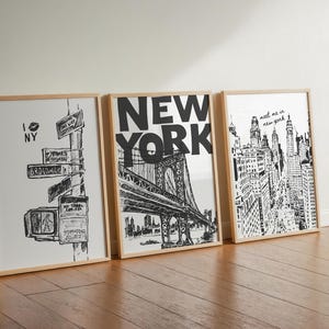May include: Three framed black and white prints leaning against a wall. The prints feature New York City landmarks and text, including a street sign, the Brooklyn Bridge, and a cityscape. The frames are light wood.