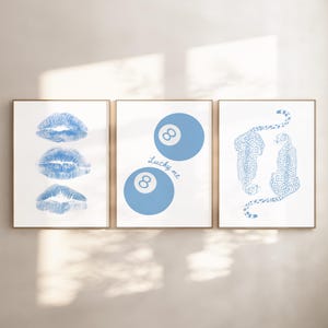 May include: Three framed art prints with gold frames. The prints feature blue designs on a white background. One print shows three blue lip imprints. Another has two blue 8-ball designs with the text "Lucky me". The third print depicts two blue cheetahs.