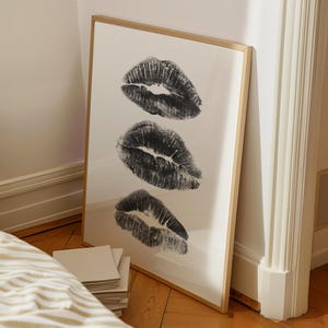 May include: A framed print with three black and white lip prints on a white background. The frame is a light wood color.