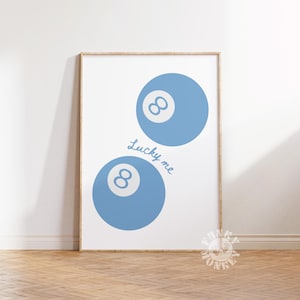 May include: A framed print featuring two light blue eight balls with the text "Lucky me" in a cursive font. The eight balls are positioned diagonally on a white background. The frame is a light wood color.