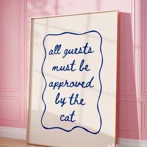 May include: A white framed poster with a blue border and the text "all guests must be approved by the cat" in blue cursive writing.