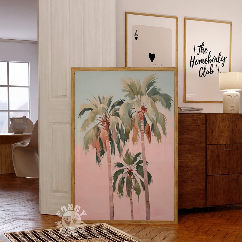 May include: A framed print of three palm trees against a pink and blue sky. The print is in a wooden frame and is hanging on a wall.