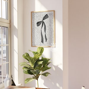 Puede incluir: Una impresi&oacute;n enmarcada de una cinta negra atada en un lazo sobre un fondo blanco con texto escrito a mano. La impresi&oacute;n est&aacute; colgada en una pared blanca junto a una planta verde.