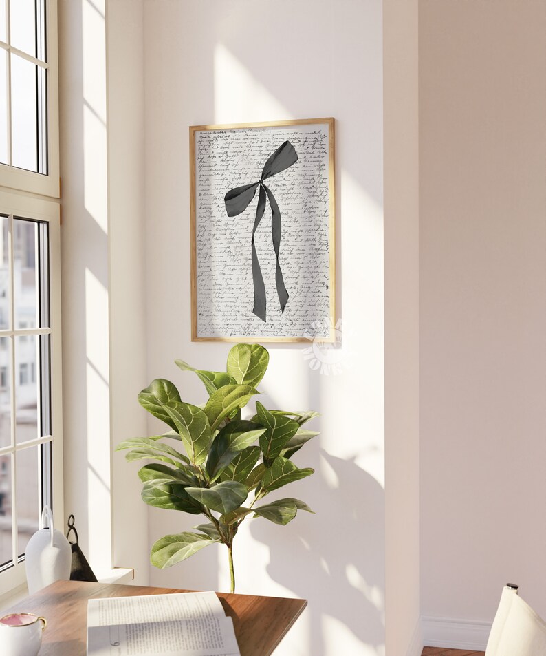 Puede incluir: Una impresi&oacute;n enmarcada de una cinta negra atada en un lazo sobre un fondo blanco con texto escrito a mano. La impresi&oacute;n est&aacute; colgada en una pared blanca junto a una planta verde.