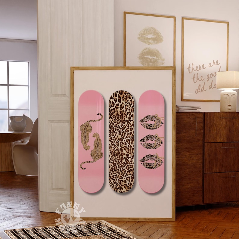 Op de afbeelding: Een ingelijst kunstwerk met drie skateboard decks. De ontwerpen omvatten roze, luipaardprint en lipmotieven. E&eacute;n deck heeft luipaarden, een ander een luipaardprint en de derde drie sets lippen.