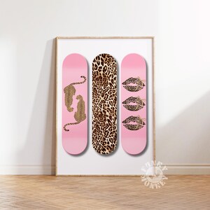 Op de afbeelding: Ingelijste kunstafdruk met drie roze skateboards. E&eacute;n heeft luipaardfiguren, een andere een luipaardprint en de derde luipaardlippen. Het kunstwerk wordt op een witte muur getoond.