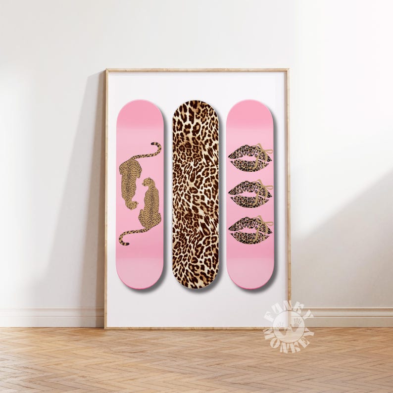 Op de afbeelding: Ingelijste kunstafdruk met drie roze skateboards. E&eacute;n heeft luipaardfiguren, een andere een luipaardprint en de derde luipaardlippen. Het kunstwerk wordt op een witte muur getoond.