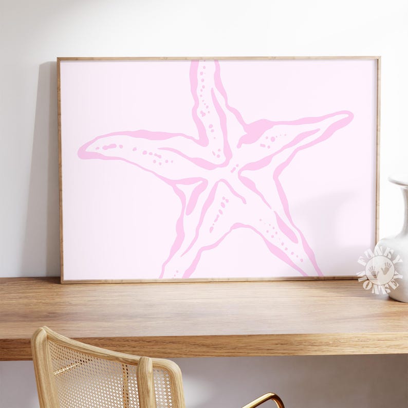 May include: A framed print featuring a pink starfish design on a light pink background. The starfish is a five-pointed star shape with a textured appearance. The frame is a light brown color, and the print is displayed on a wooden surface.