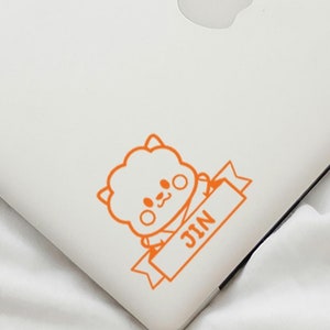 May include: A white laptop with an orange sticker of a cartoon character with the word "JIN" on a banner.