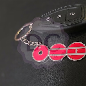 May include: A car key and a keychain with a red and silver emblem. The keychain has a series of red circles with silver outlines and a silver chain. The car key is black.