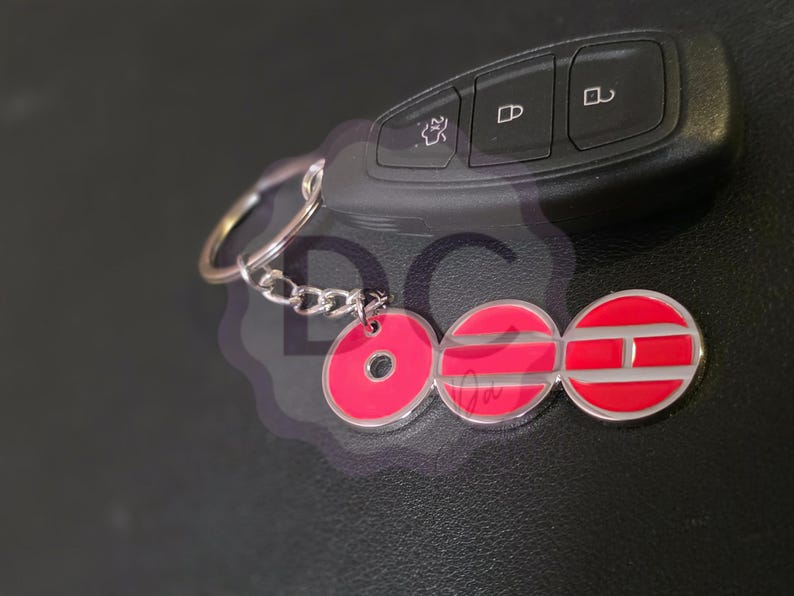 May include: A car key and a keychain with a red and silver emblem. The keychain has a series of red circles with silver outlines and a silver chain. The car key is black.