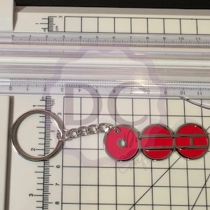 May include: A silver keychain with a circular key ring and chain, connected to three red, circular charms. The charms have a silver border. The keychain is on a white surface with a ruler showing inches and centimeters.