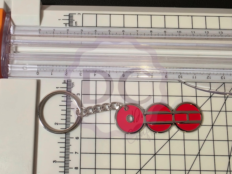 May include: A silver keychain with a circular key ring and chain, connected to three red, circular charms. The charms have a silver border. The keychain is on a white surface with a ruler showing inches and centimeters.