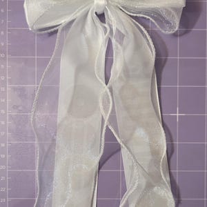 May include: A white hair bow made of sheer, translucent organza ribbon with a metallic alligator clip attached to the center back. The bow features two long, flowing tails and is positioned against a purple cutting mat with white grid lines.