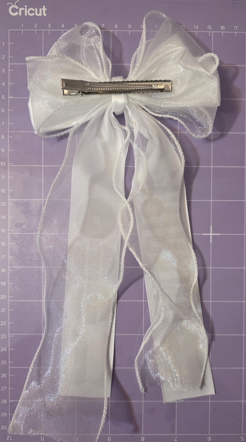May include: A white hair bow made of sheer, translucent organza ribbon with a metallic alligator clip attached to the center back. The bow features two long, flowing tails and is positioned against a purple cutting mat with white grid lines.
