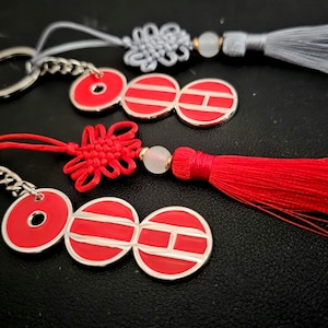 May include: Two keyrings with red and white circular charms and silver key rings. One keyring has a red tassel and knot, the other has a grey tassel and knot. The charms have a coin-like design.