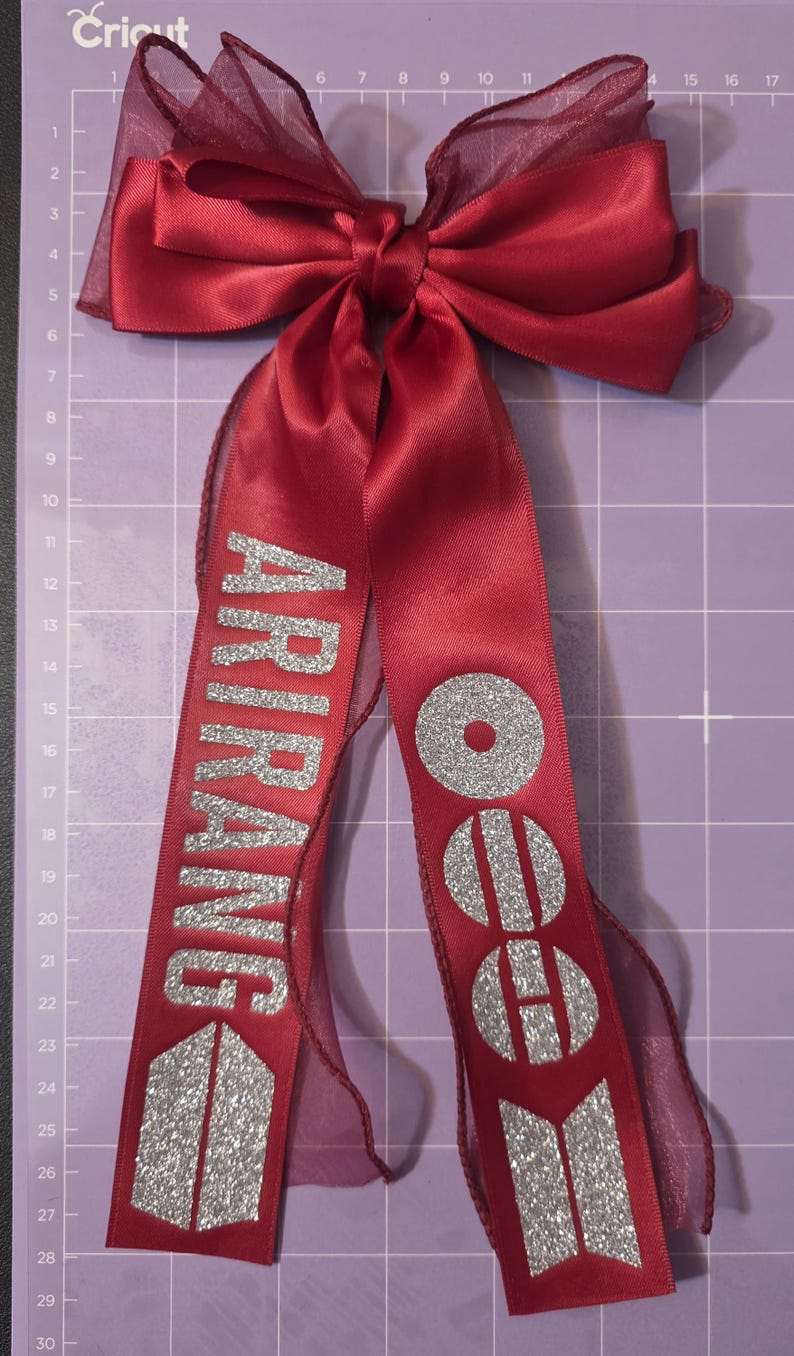 May include: A deep red satin hair bow featuring two long, dangling ribbons. The left ribbon displays the silver glitter text ARIRANG, and the right ribbon features a silver glitter BTS logo. The bow is layered with a sheer, dark red organza fabric.