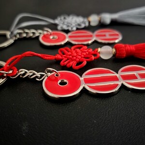 May include: Three keychains with red and silver accents. Each keychain features a silver ring and chain, connected to a red charm with a silver border. One has a red tassel, another a gray tassel, and the third a red knot.