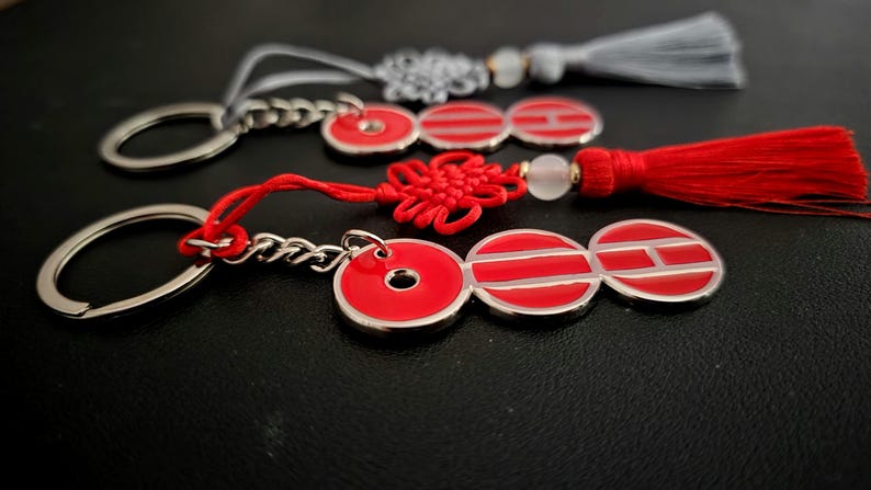 May include: Three keychains with red and silver accents. Each keychain features a silver ring and chain, connected to a red charm with a silver border. One has a red tassel, another a gray tassel, and the third a red knot.