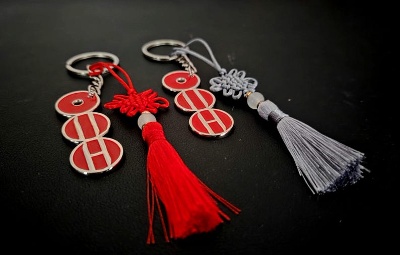 May include: Keychains with silver rings and red and white circular pendants. One keychain has a red tassel, another has a red knot, and the third has a gray tassel. The pendants have the letter "H" on them.