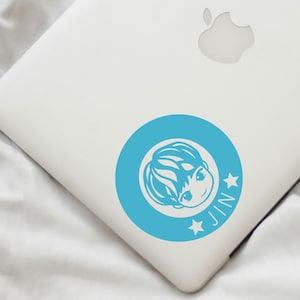 May include: A blue circle sticker with a cartoon drawing of a person's head inside. The person has short, dark hair and is smiling. Two white stars are on either side of the word "JIN" in white letters.