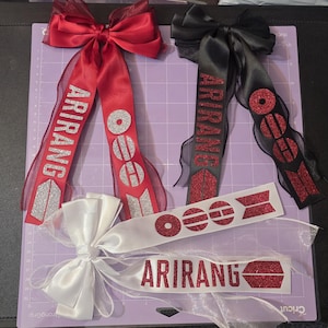 Può includere: Tre fiocchi decorativi per capelli in nastro di raso rosso, nero e bianco. Ogni fiocco presenta la scritta glitterata ARIRANG e un logo circolare con linee orizzontali. Il fiocco rosso ha glitter argento, quello nero e quello bianco hanno glitter rossi.