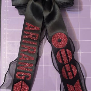 May include: A black satin hair bow featuring two long, dangling ribbon tails. The left tail displays the red glitter text ARIRANG above a red glitter logo, while the right tail features red glitter circles and a red glitter geometric symbol.