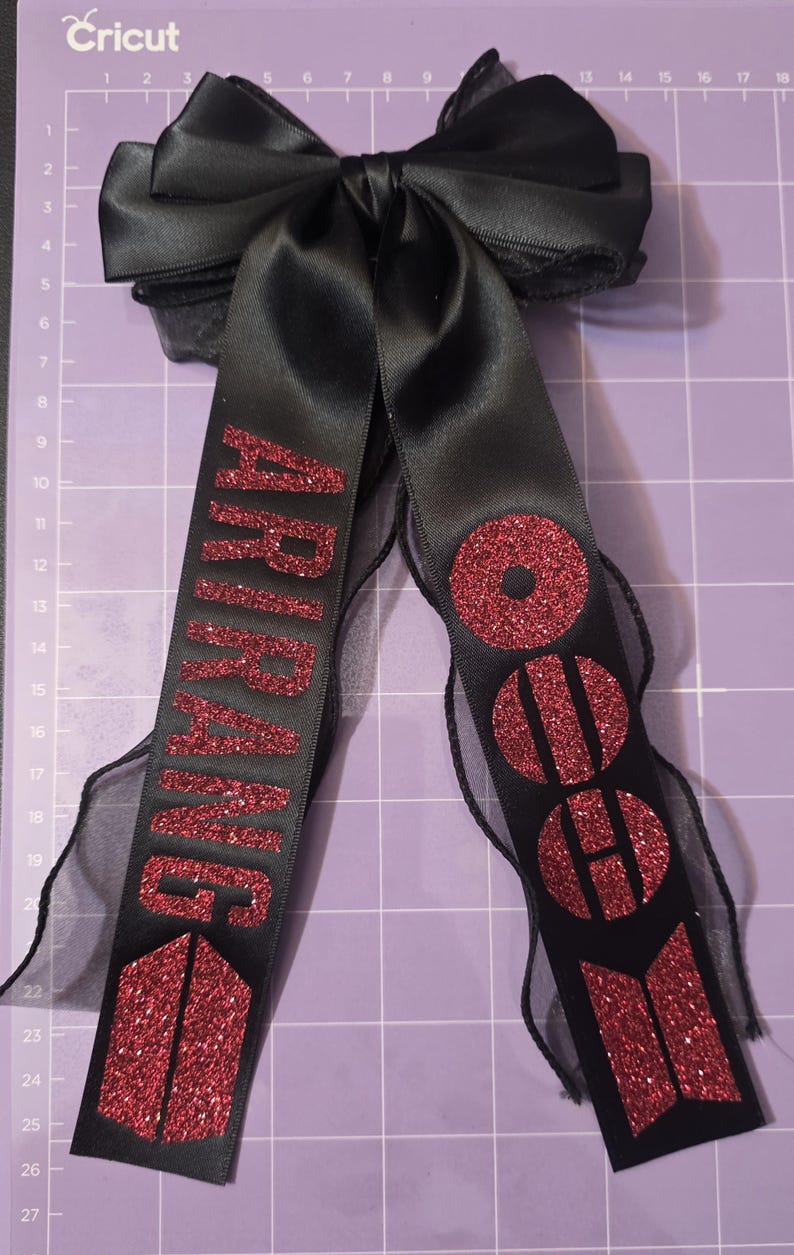 May include: A black satin hair bow featuring two long, dangling ribbon tails. The left tail displays the red glitter text ARIRANG above a red glitter logo, while the right tail features red glitter circles and a red glitter geometric symbol.