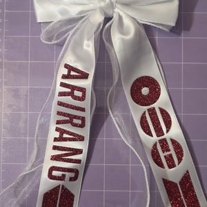 May include: A white satin and sheer ribbon bow featuring two long tails. The left tail displays the word ARIRANG in dark red glitter, and the right tail features three circular dark red glitter icons with a horizontal line through each.