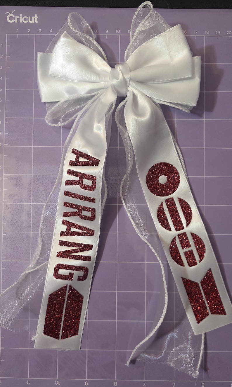 May include: A white satin and sheer ribbon bow featuring two long tails. The left tail displays the word ARIRANG in dark red glitter, and the right tail features three circular dark red glitter icons with a horizontal line through each.