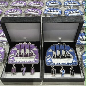 May include: Two black display boxes containing enamel pins shaped like ships with sails. One pin features purple tones, the other blue. Each ship has three dangling charms: a logo, the number 7, and a geometric shape. The ship hulls display the text "Eternity".