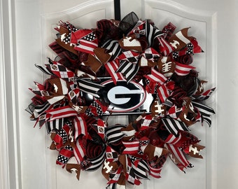 May include: A red, black, and white wreath with a metal sign featuring a 'G' in the center. The wreath is decorated with ribbons, mesh, and football-themed accents.