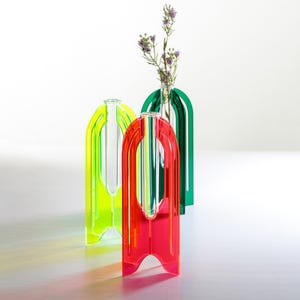 Op de afbeelding: Drie kleurrijke, moderne vazen in neongeel, rood en groen. Elke vaas heeft een boogvormig ontwerp met een centrale opening. De groene vaas bevat een klein boeket paarse bloemen. De vazen zijn gemaakt van een doorschijnend materiaal.