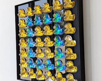 May include: A framed artwork featuring a grid of yellow and iridescent blue glass duck figurines. The ducks are arranged in rows and columns against a black and silver background, encased in a black frame. The artwork is mounted on a white wall.