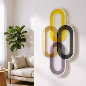 May include: Abstract wall art featuring interlocking oval shapes in yellow, gold, brown, dark green, and purple. The artwork is encased in a white frame and mounted on a white wall. A living room setting with a plant and sofa is visible.