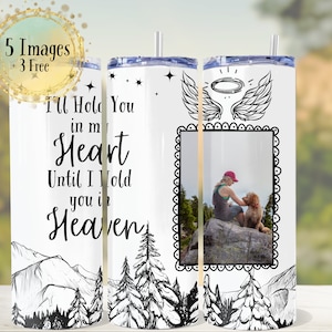 May include: White stainless steel tumbler with a black and white drawing of a mountain range, trees, and a pair of angel wings. The tumbler has a black and white photo frame with a picture of a person and a dog sitting on a rock. The text "I'll Hold You in My Heart Until I Hold You in Heaven" is printed on the tumbler.