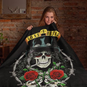 Puede incluir: Manta negra con el logotipo de Guns N' Roses, una calavera con sombrero, pistolas cruzadas y rosas rojas. El nombre de la banda se muestra en letras doradas sobre una pancarta amarilla. El diseño está sobre un fondo negro.