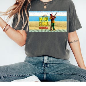 May include: A gray t-shirt with a vintage-style graphic of a man playing a guitar on a beach. The graphic includes the text "Manu Chao Esperanza".
