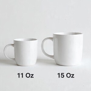 Op de afbeelding: Twee effen witte keramische mokken met oren. De kleinere mok links is gelabeld met 11 Oz, en de grotere mok rechts is gelabeld met 15 Oz. Beide mokken hebben een gladde, glanzende afwerking en een eenvoudig, afgerond ontwerp voor warme dranken.