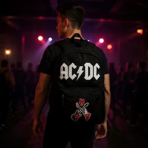 May include: Black backpack featuring the AC/DC logo in white, with a lightning bolt. Below the logo is a patch with two crossed guitars and the text "Rock and Roll". The backpack is worn by a person in a black t-shirt.